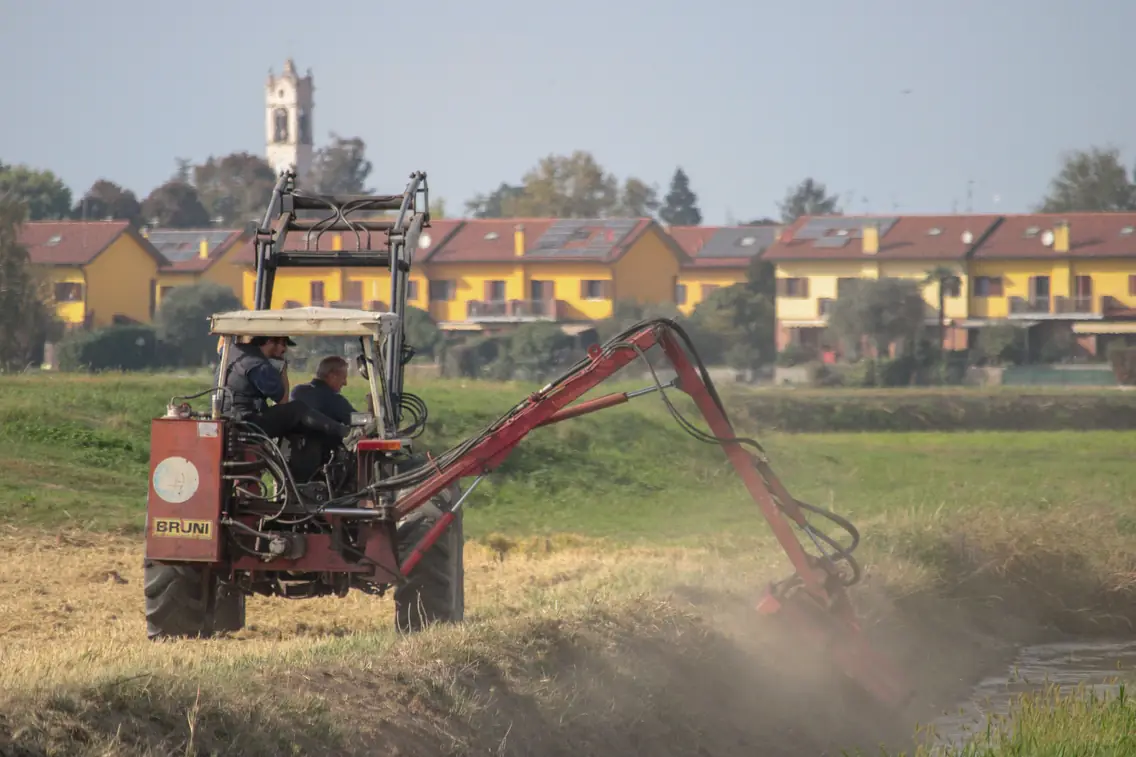 Tracteur équipé d’un bras de débroussaillage entretenant un fossé, village en arrière-plan